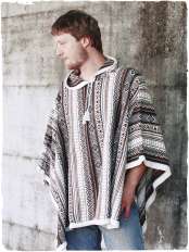 Mann in Jeans, Hände in den Taschen vor einer Wand, trägt rechteckigen Alpaka-Poncho, weiße Basis und Naturtöne, Kapuze, Modell Mexico.