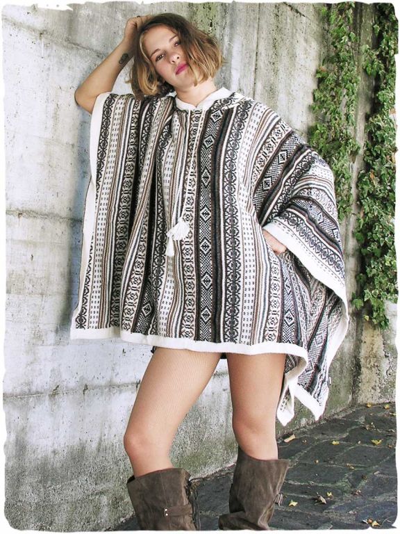 Frau vor heller Wand mit Efeu, trägt einen rechteckigen Poncho aus Alpakawolle in natürlichen Ethno-Tönen, mit Kapuze, Modell Mexico.