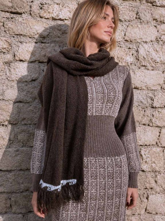 Blonde Frau vor einer Steinmauer mit Strickkleid und braunem Damen-Winterschal aus Alpakawolle mit Fransen, Modell Kora.